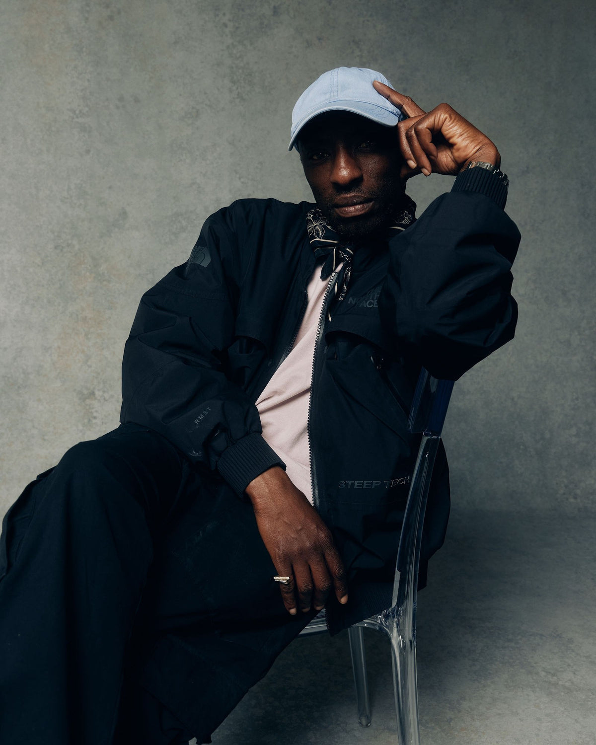 Walé Adeyemi seated in a grey studio, pale blue cap, hand raised to the brim